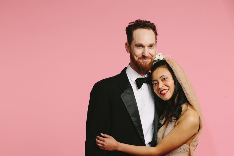 A smiling portrait of a bride and groom embraced, isolated on pink background